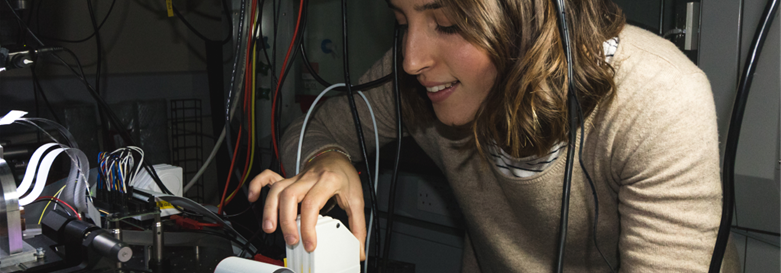 Smiling female student examines and adjusts quantum engineering equipment. 